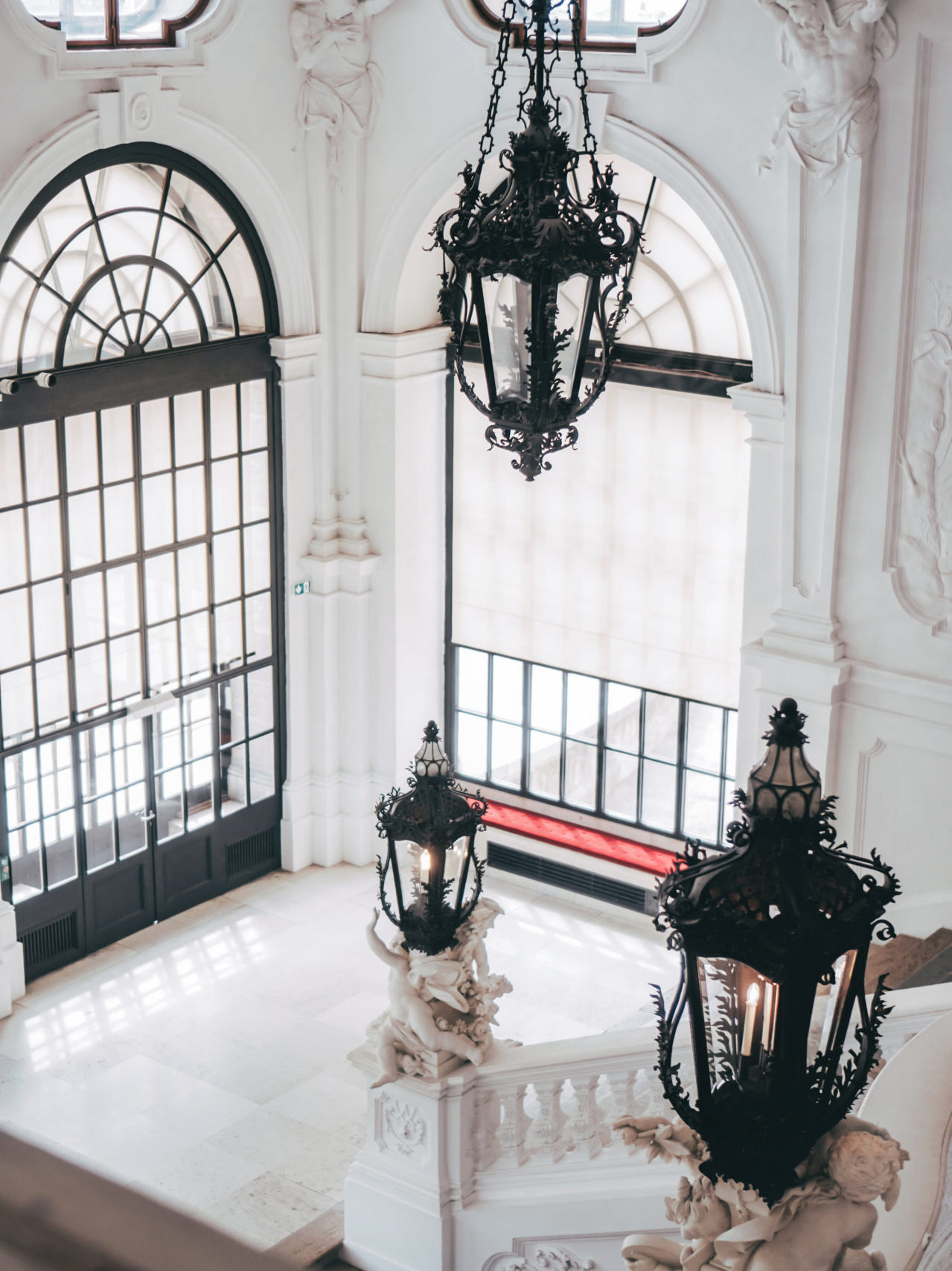 Vienna Travel Guide: A white baroque staircase decorated with statues holding ornate black lanterns in Belevedere Museum in Austria