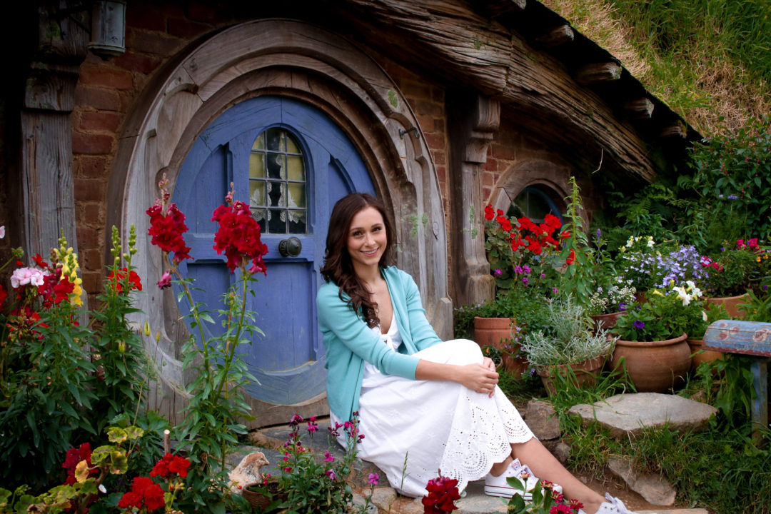 Travel Blogger Jordan Gassner sitting in front of a hobbit hole with a blue door in Hobbiton New Zealand