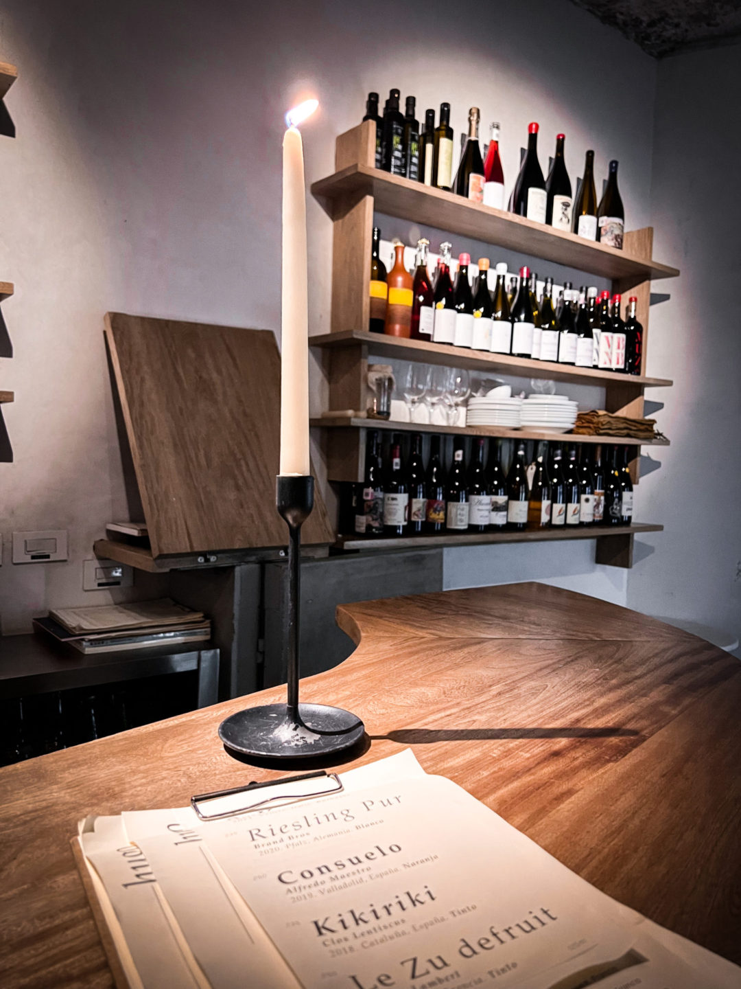 A lit candle and a wine list in front of a shelf with multiple bottles of natural, organic and biodymanic wines at Hugo Wine Bar in Mexico City