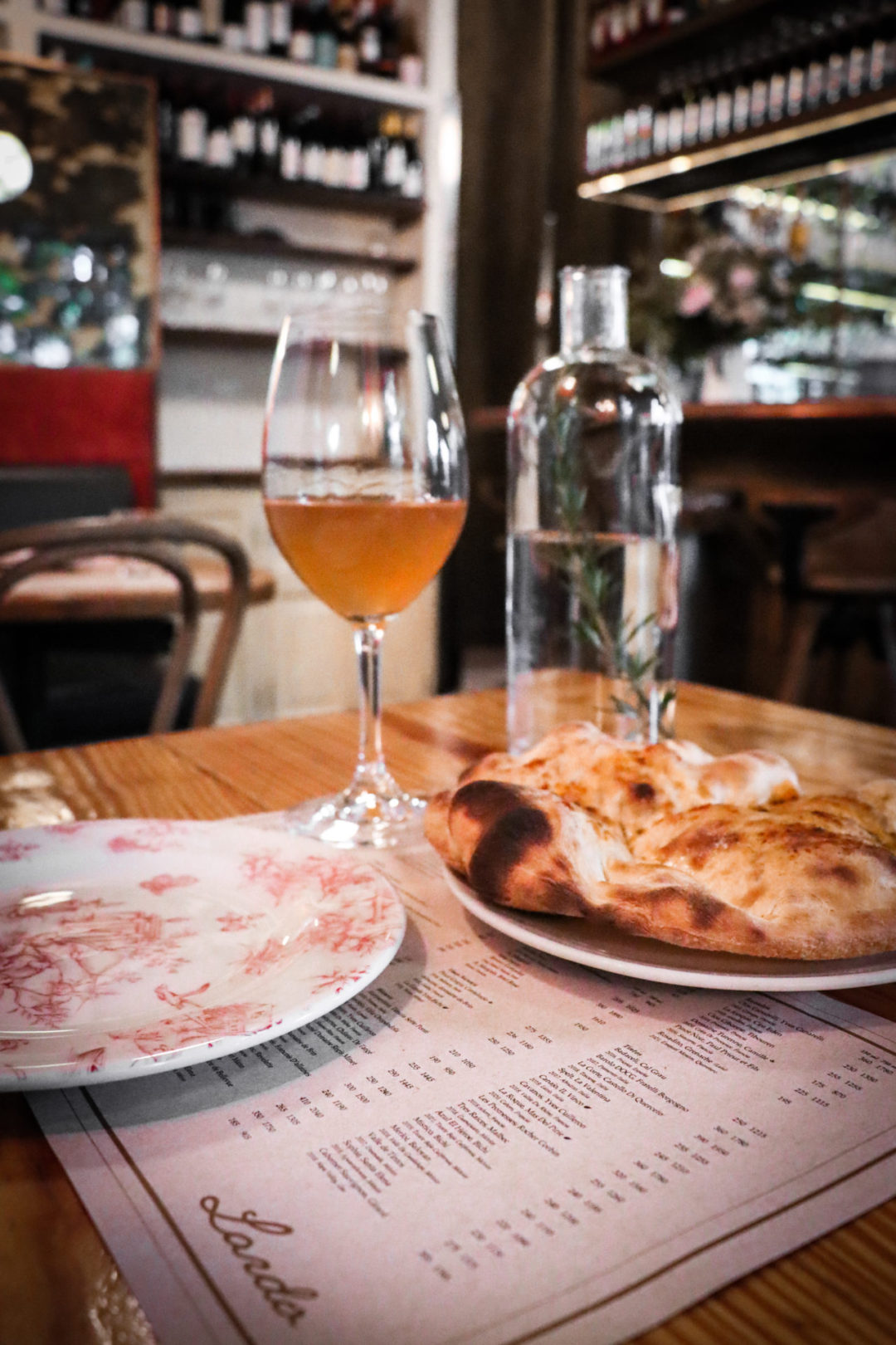 A glass of orange wine, a serving tray with flatbread and a red and white china plate sit on top of a Lardo Restaurant menu in CDMX