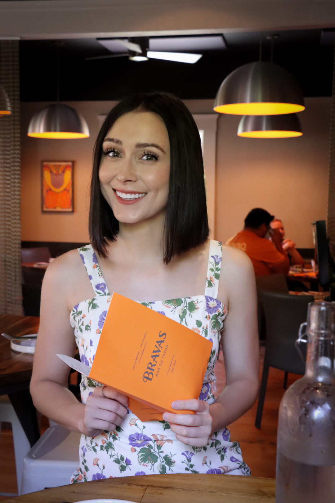 Healdsburg Travel Guide: Travel Blogger Jordan Gassner wearing a white floral dress and seated inside smiling while holding a "Bravas Bar De Tapas" Restaurant Menu