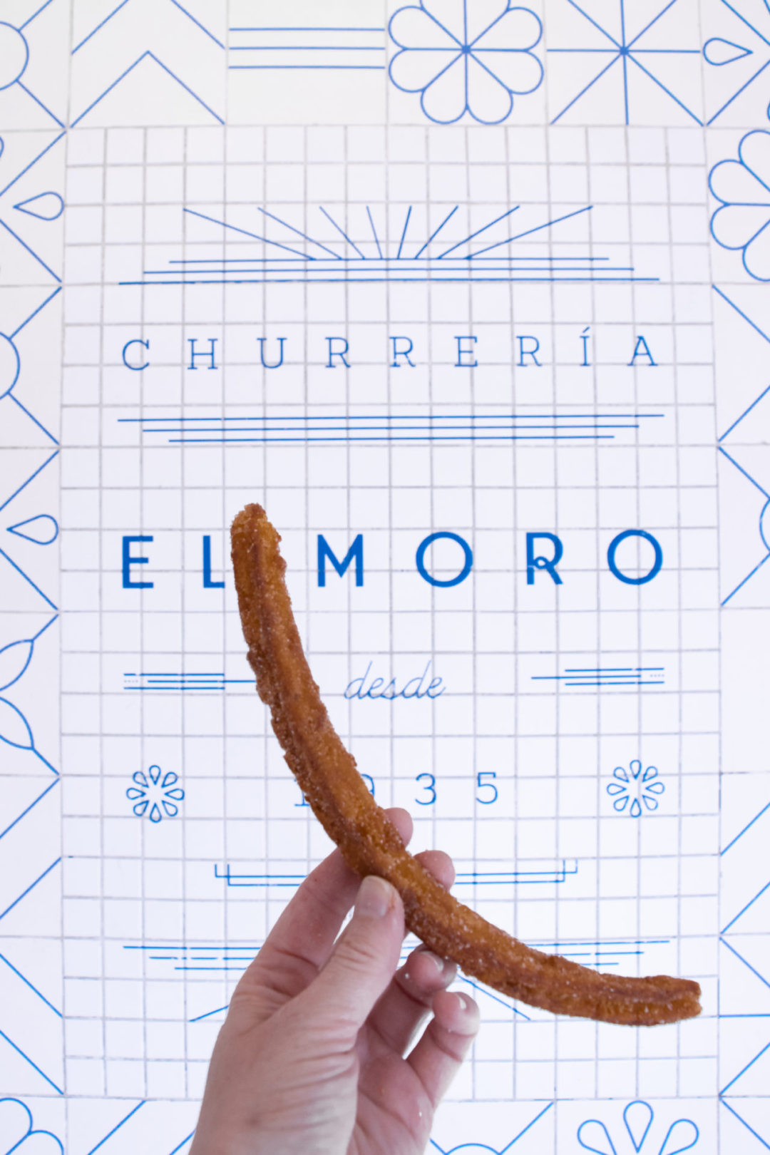 Dairy-free in Mexico City: Travel Blogger Jordan Gassner holding up a solo churro in front of a blue and white Churrería El Moro sign.
