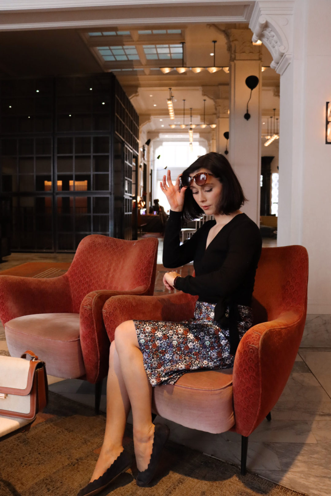Travel Blogger Jordan Gassner looking at her watch while sitting on a burnt orange accent chair inside the Kelly Wearstler designed San Francisco Proper Hotel Lobby