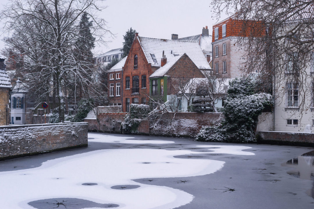 A frozen canal covered in snow in Bruges, Belgium snaking toward its left with traditional colorful houses dotting either side