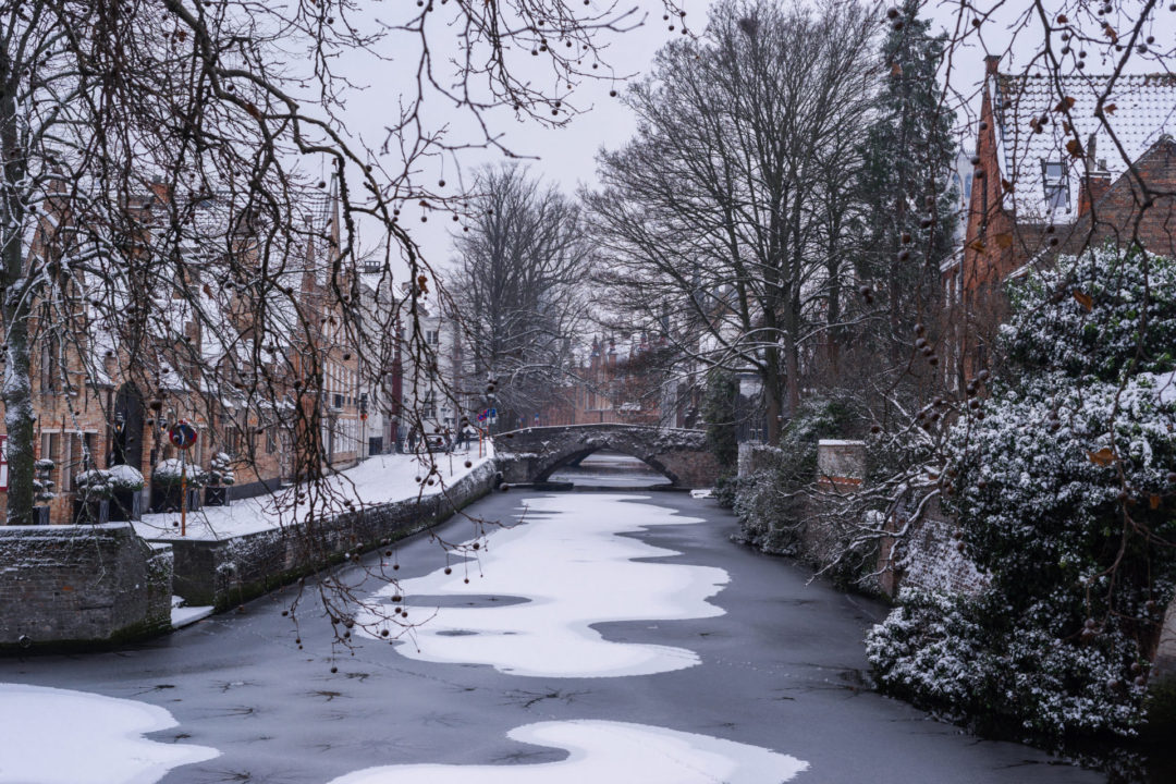 A straight, frozen canal covered with a layer of snow and with traditional homes and shops on either side in Bruges, Belgium