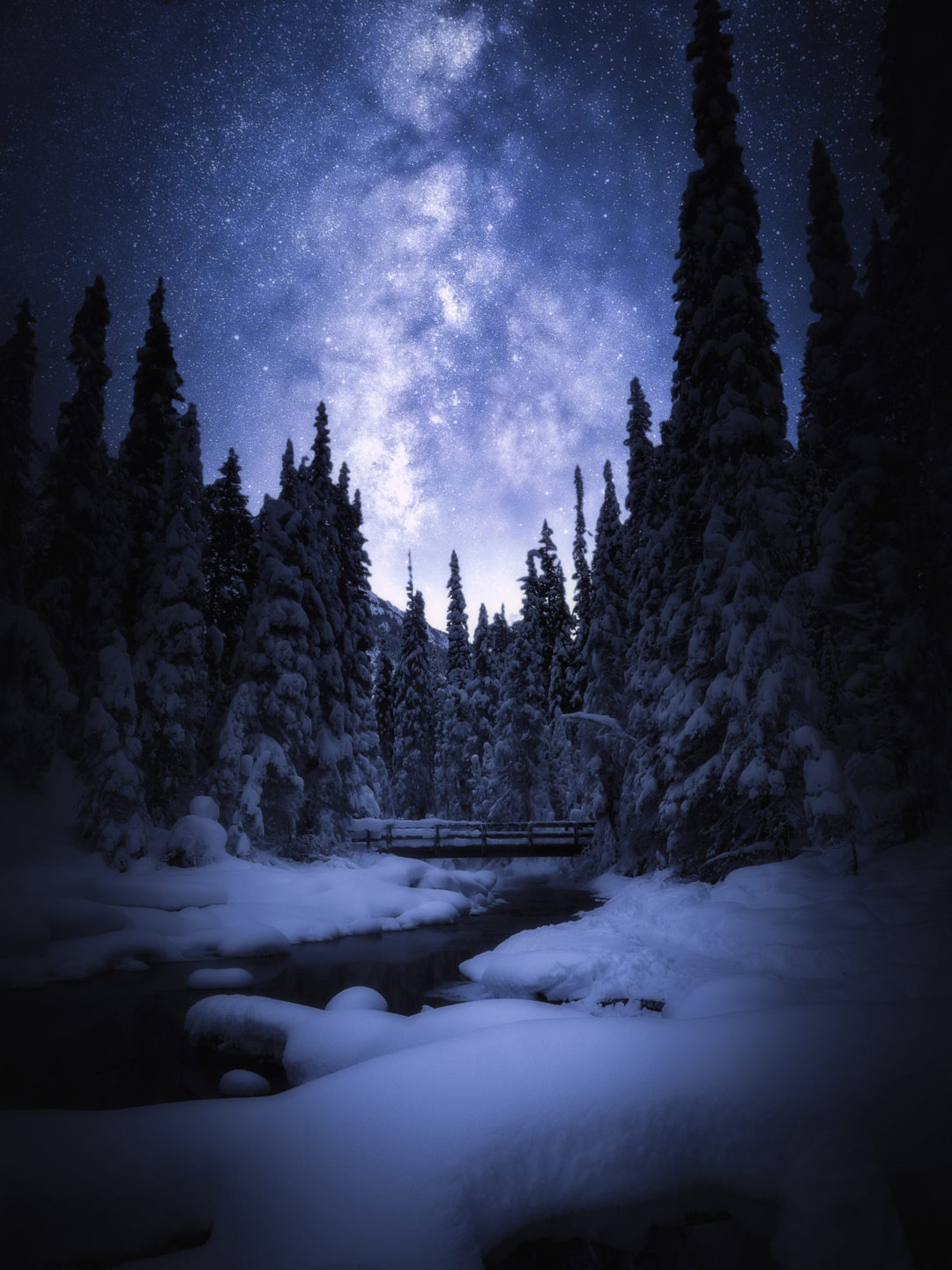 A clear night sky under the snowy landscape of a wooden bridge spanning a river in a forest