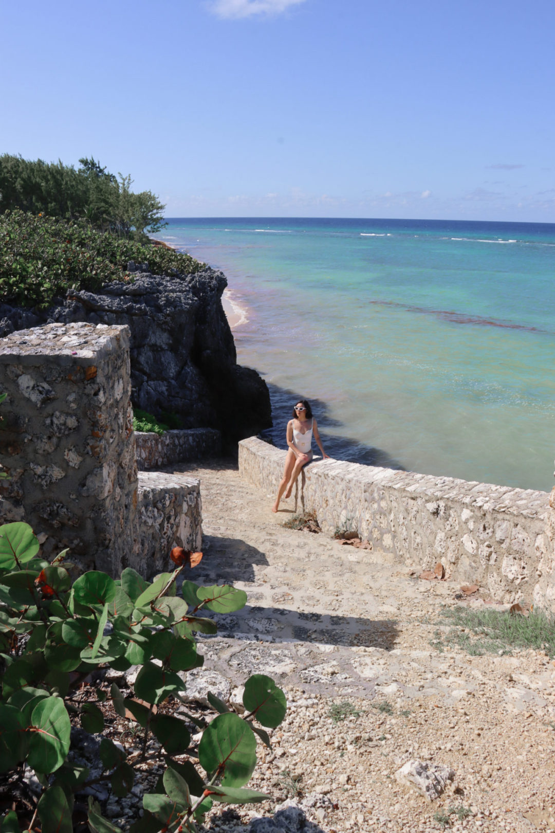 Travel Blogger Jordan Gassner wearing a white one piece swimsuit and sunglasses at the Barefoot Beach staircase in the Cayman Islands