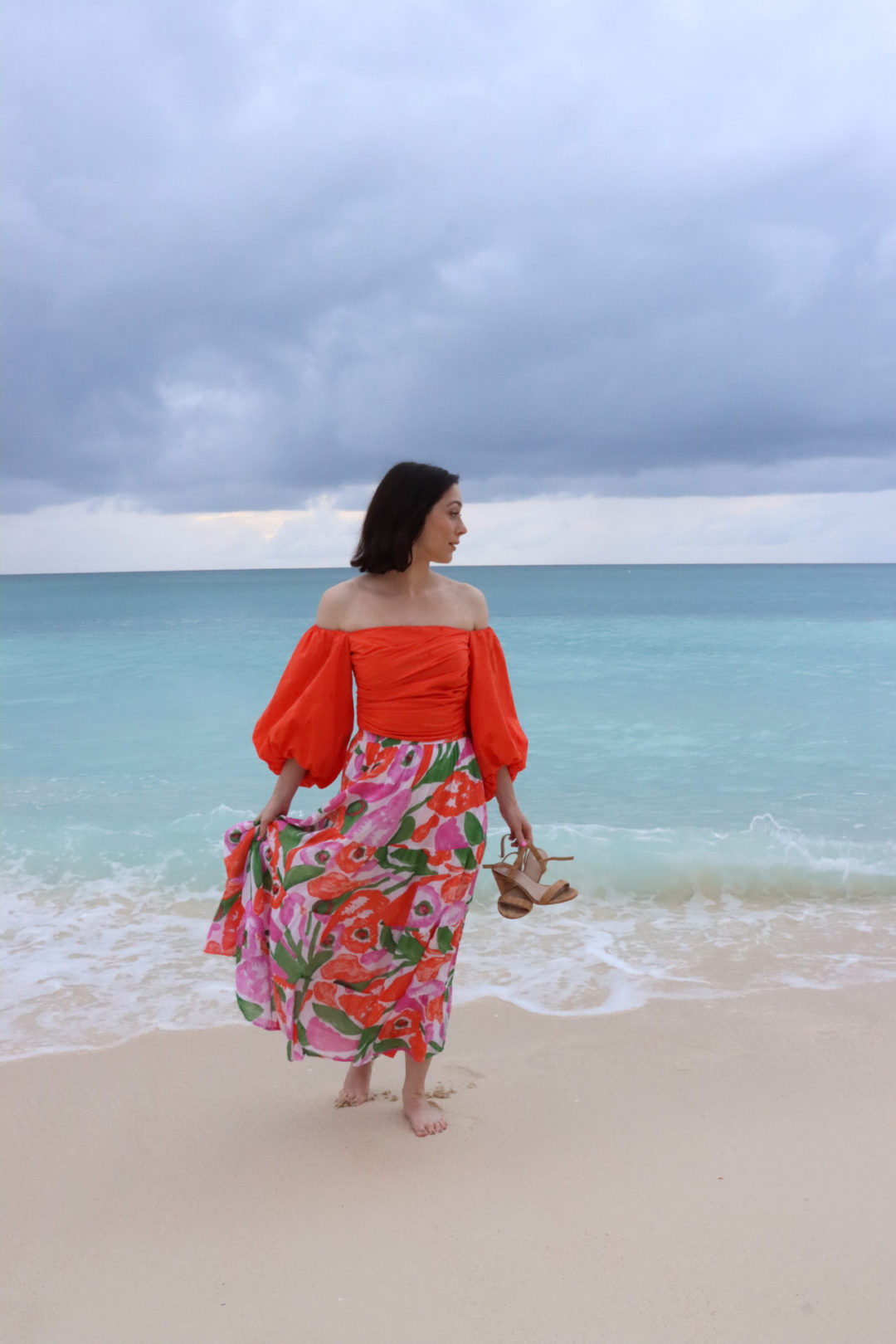 Travel Blogger Jordan Gassner holding a pair of heels and her skirt while standing near a turquoise blue shoreline on Grand Cayman Island