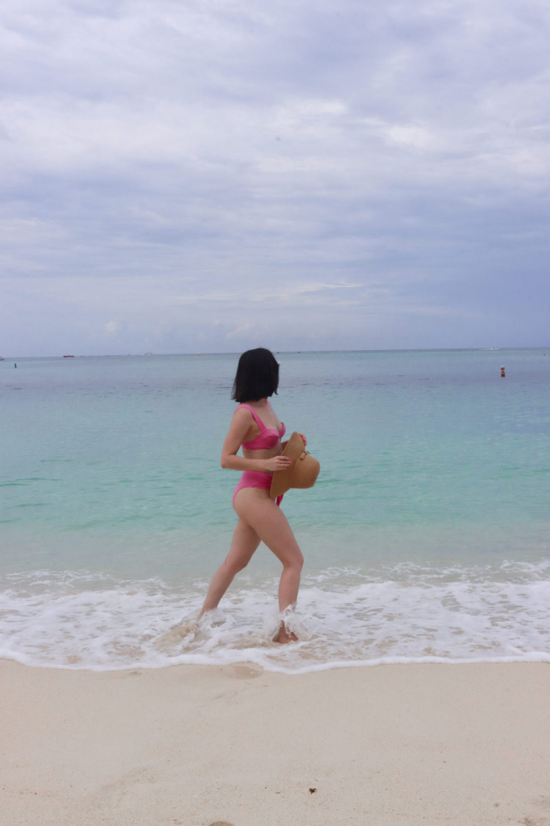Travel Blogger Jordan Gassner holding a straw hat while walking the Governor's Beach shoreline on Grand Cayman Island in the Caribbean