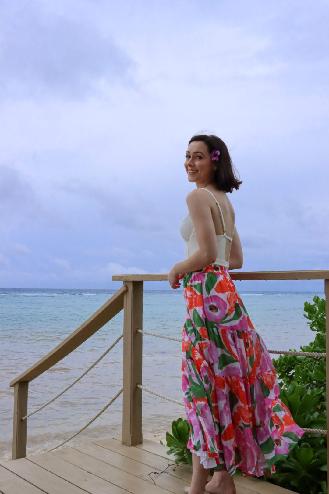 Travel Blogger Jordan Gassner standing on a dock and smiling in a white bathing suit and floral print skirt on the North Side of Cayman Islands