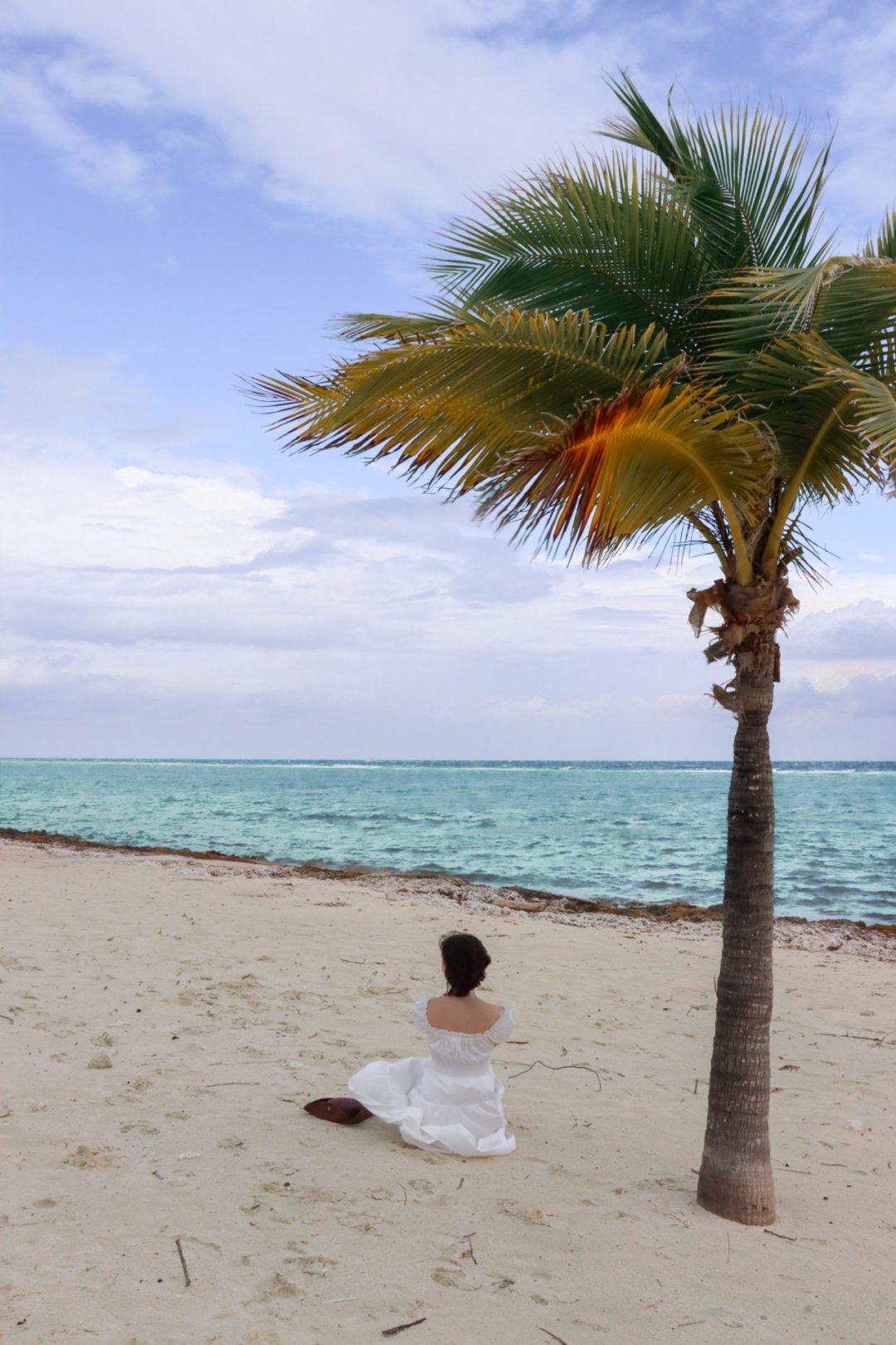 Travel Blogger Jordan Gassner wearing a white dress and sitting next to a pirate's hat and under a palm tree at Rum Point in Grand Cayman