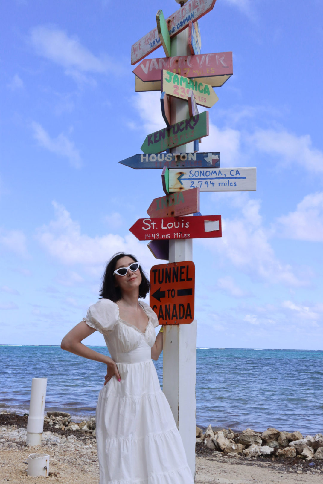 Travel Blogger Jordan Gassner standing near a colorful sign post at Tukka Restaurant on Grand Cayman marking the distances to other famous points in the world