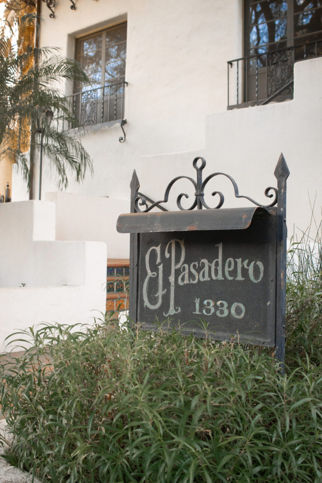A iron sign reading "El Pasadero 1330" in vintage script in front of a Spanish Colonia Revival Property in West Hollywood, California