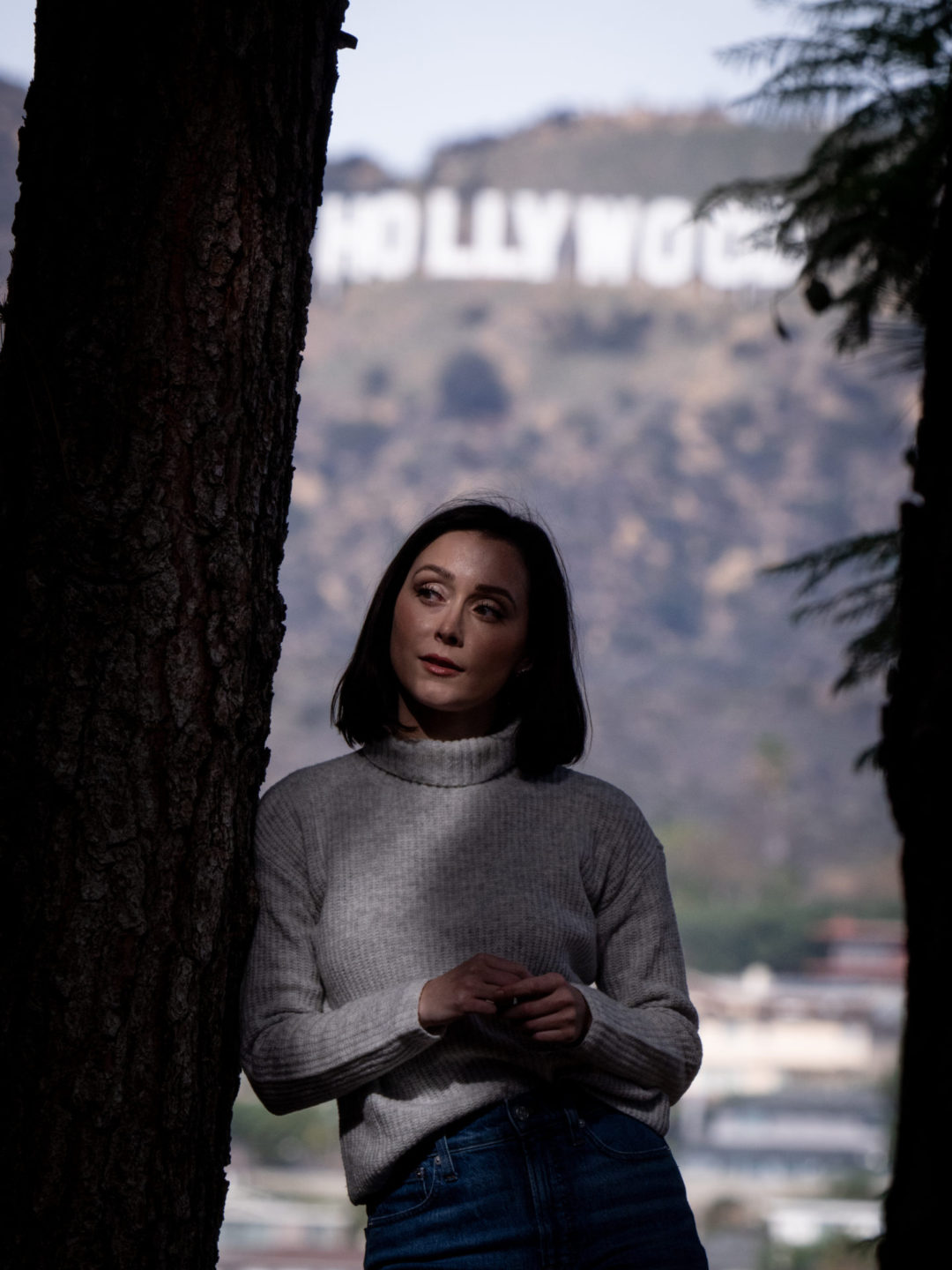 Travel Blogger Jordan Gassner peering to the side while leaning against a tree at the Los Angeles Barnsdall Art Park with the Hollywood Sign in the background