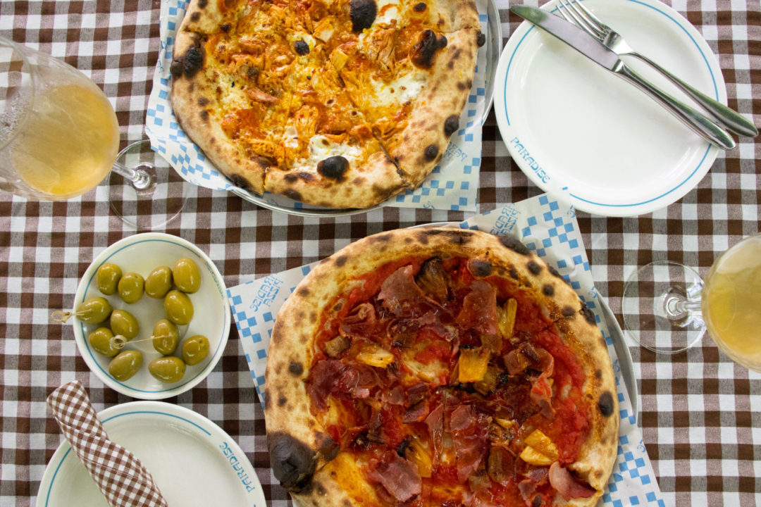 Paradise Pizza's La Hawaiiana and The New Yorker Pies on a brown and white checked tablecloth with olives and glasses of orange wine