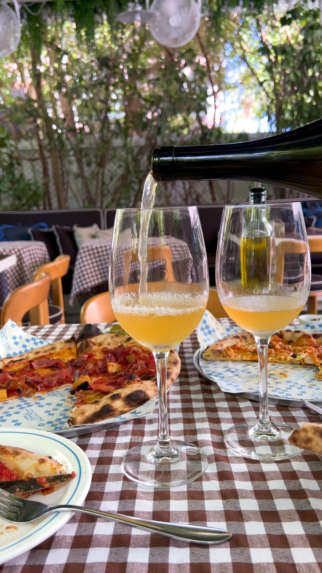 A bottle of Moby Dick Orange Wine being poured into two wine glasses next to two half-eaten pizzas with "Paradise Pizza" branded paper