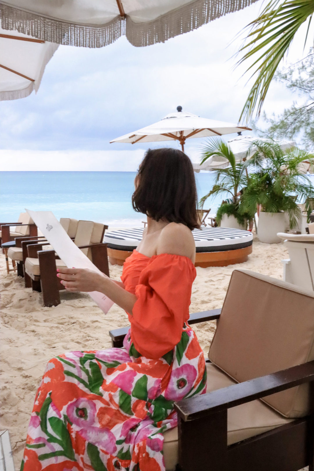 Travel Blogger Jordan Gassner looking toward the ocean while sitting in a chair on the beach with a Tillie's Restaurant Menu in Grand Cayman