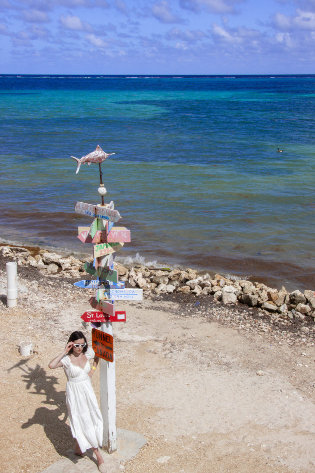 Travel Blogger Jordan Gassner adjusting her sunglasses next to a colorful directional sign-post on the east end beach of Grand Cayman
