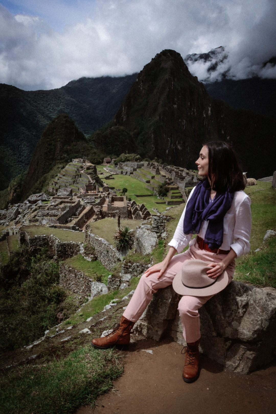 Travel Blogger Jordan Gassner taking her hat off and smiling while looking down over Machu Picchu on a semi-cloudy afternoon day