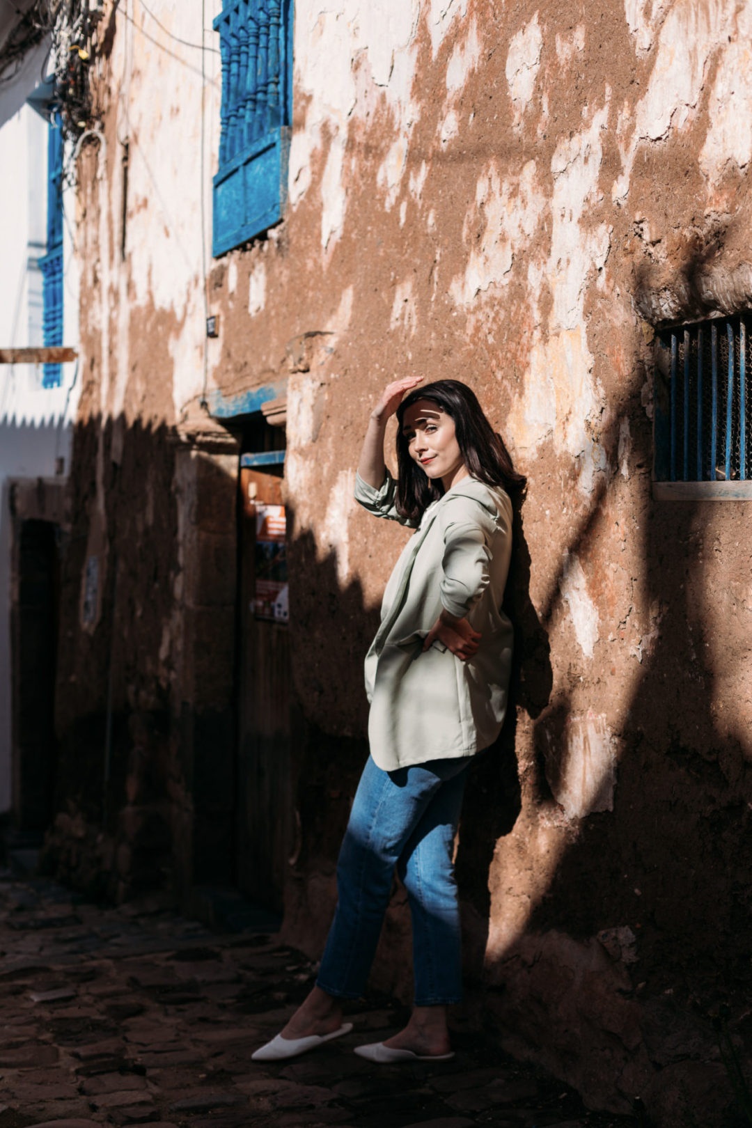 Travel Blogger Jordan Gassner shielding her eyes while standing in half sun and half shadow near a brown, white and blue building in Cusco