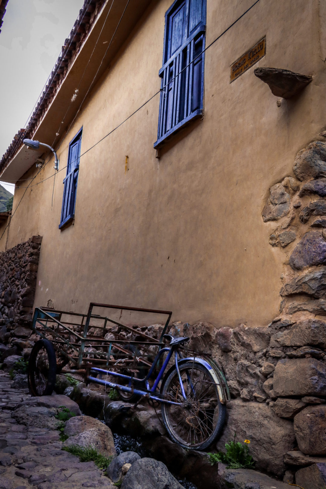 A blue tricycle and cart parked alongside a beige stucco building with a stone bottom and blue balconies