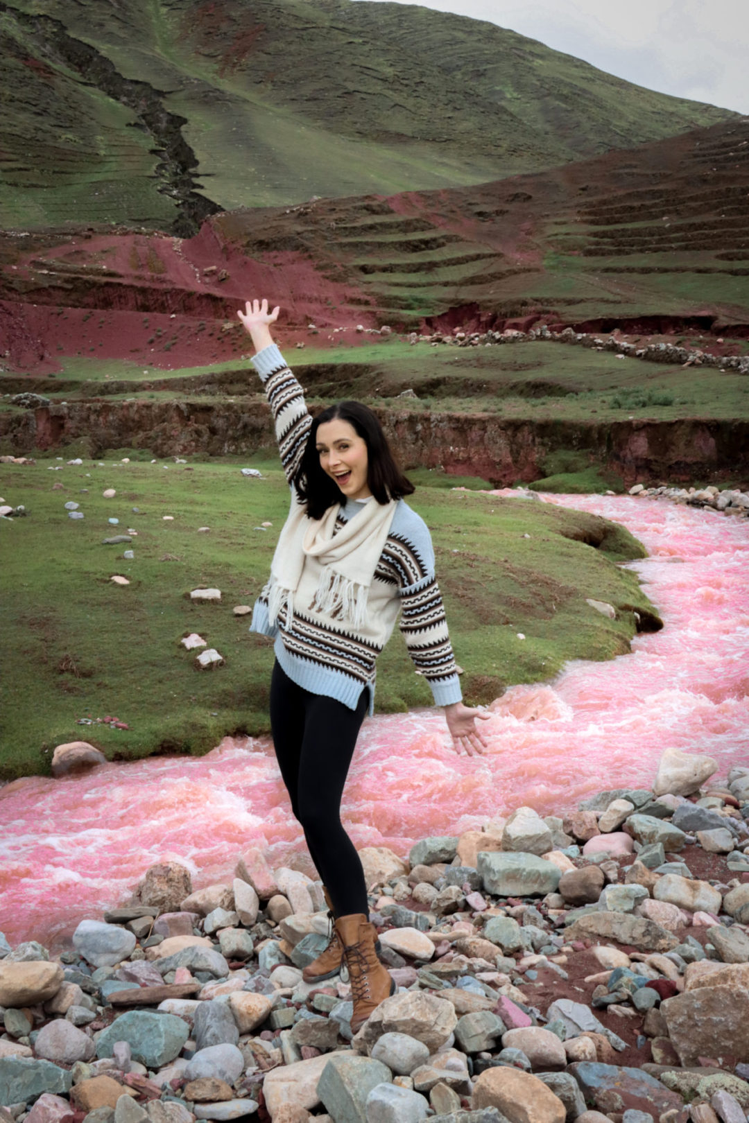 Travel Blogger Jordan Gassner smiling with hands in the air in front of the naturally pink river in Palccoyo, Peru