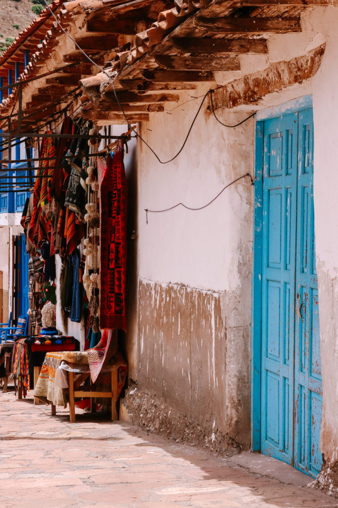 A light blue painted set of doors next to a shop selling Andean wool and clothing in Pisac, Peru's Plaza De Armas
