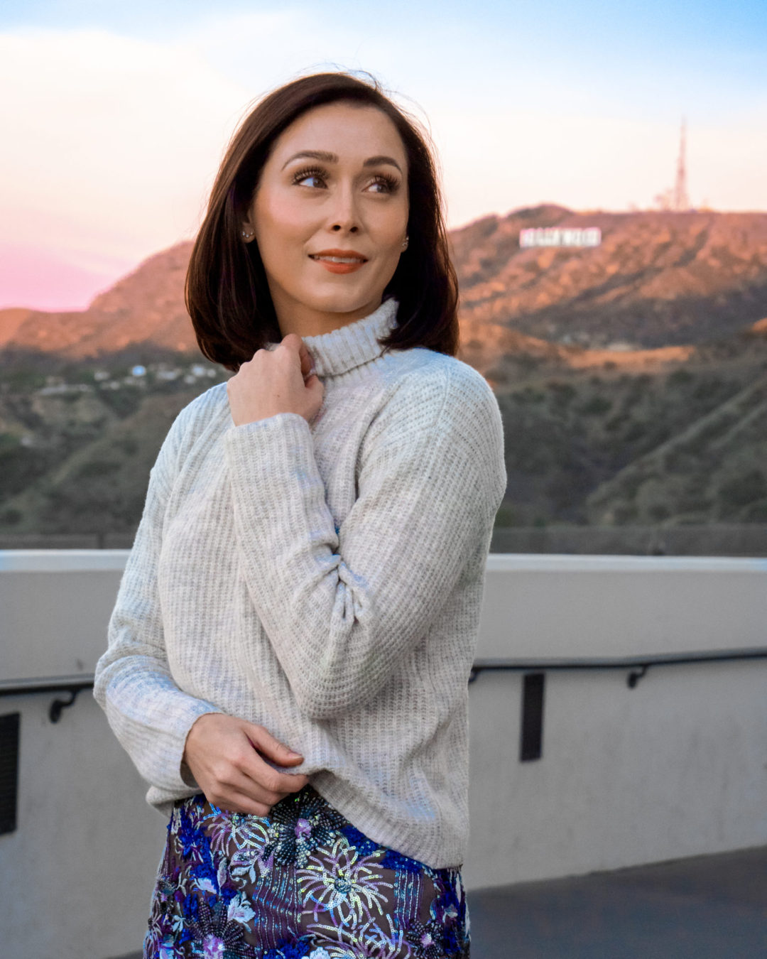 Travel Blogger Jordan Gassner bundled up in a grey sweater and blue sparkle skirt at Griffith Observatory with the Hollywood Sign in the background