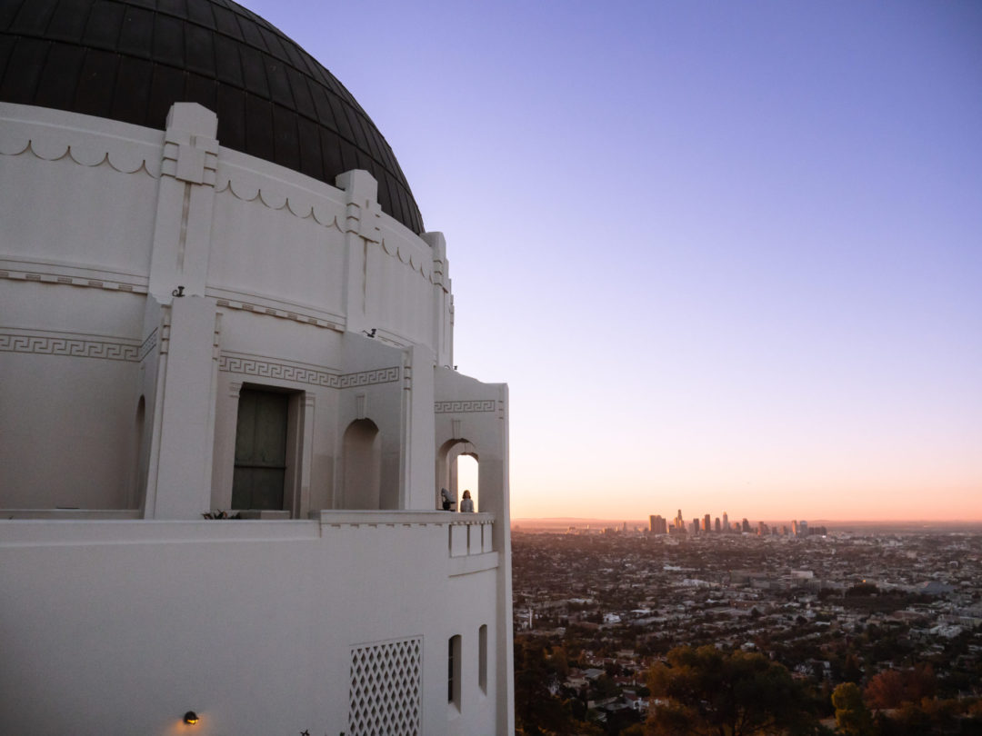 Travel Blogger Jordan Gassner standing under a wide shot of the Griffith Observatory archway overlooking Los Angeles at sunrise