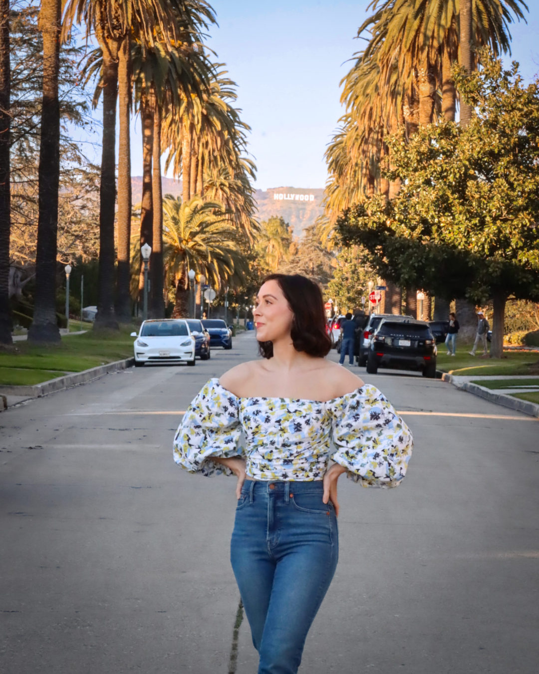 Travel Blogger Jordan Gassner standing with hands on hips and smiling to the side on a palm-tree lined street in front of the Hollywood Sign