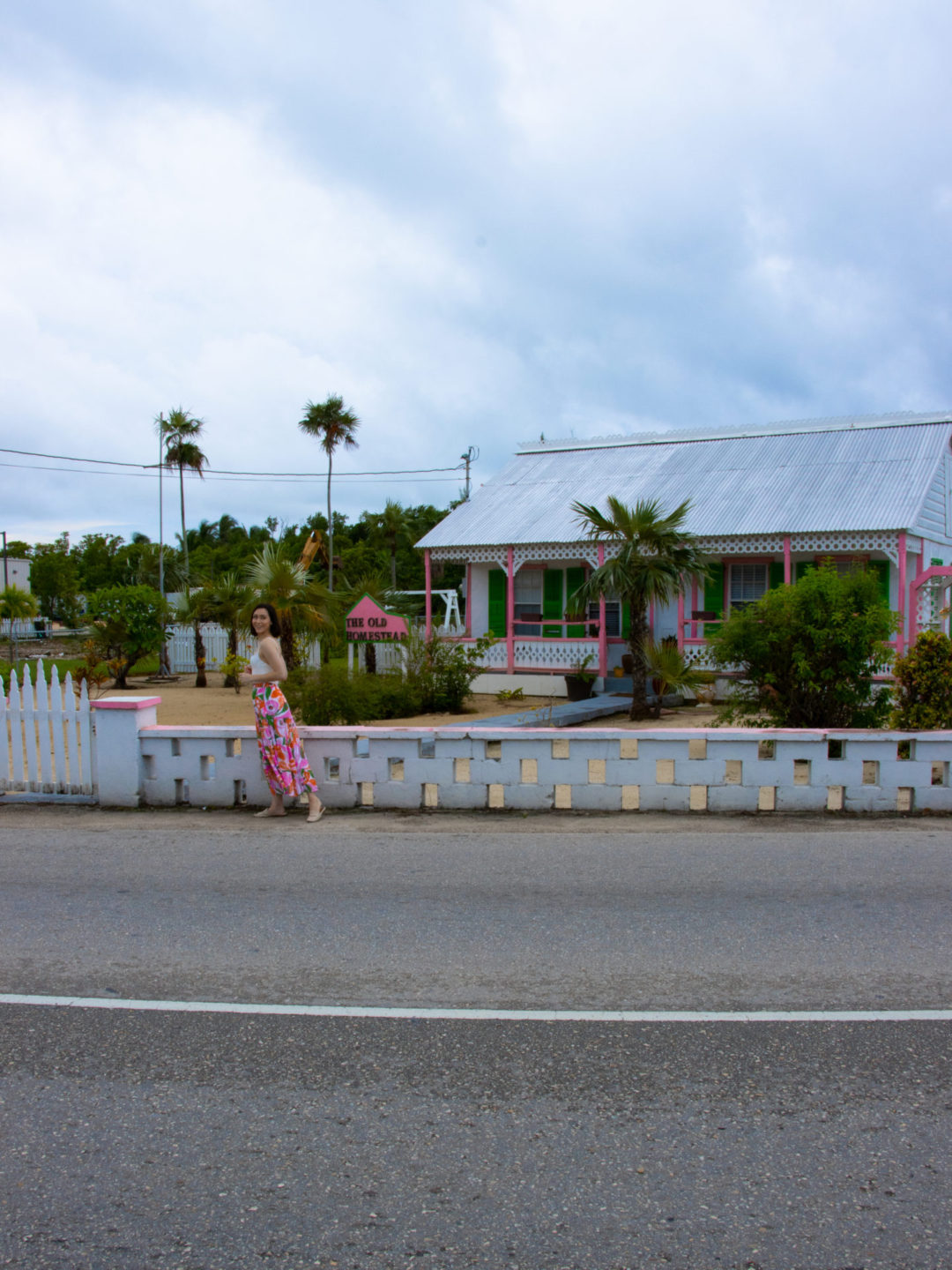 Travel Blogger Jordan Gassner smiling over her shoulder in front of a white, pink and green traditional Cayman Islands home