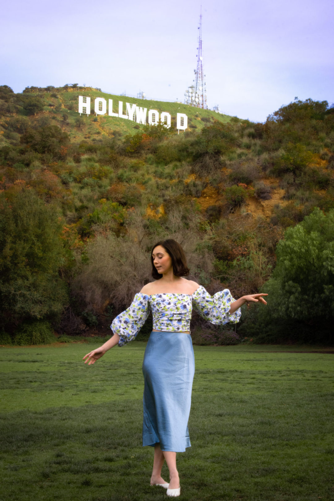 Travel Blogger Jordan Gassner looking down in a princess-style pose while snapping Hollywood Sign photos at Lake Hollywood Park