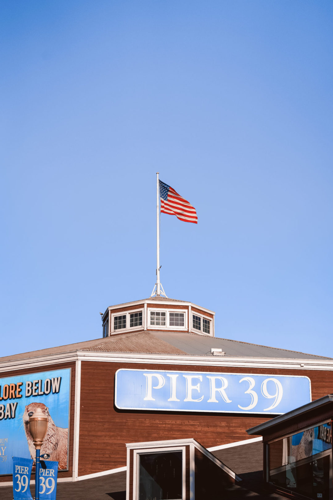 An American flag flying over a wooden building with a blue and white "Pier 39" sign in San Francisco, California