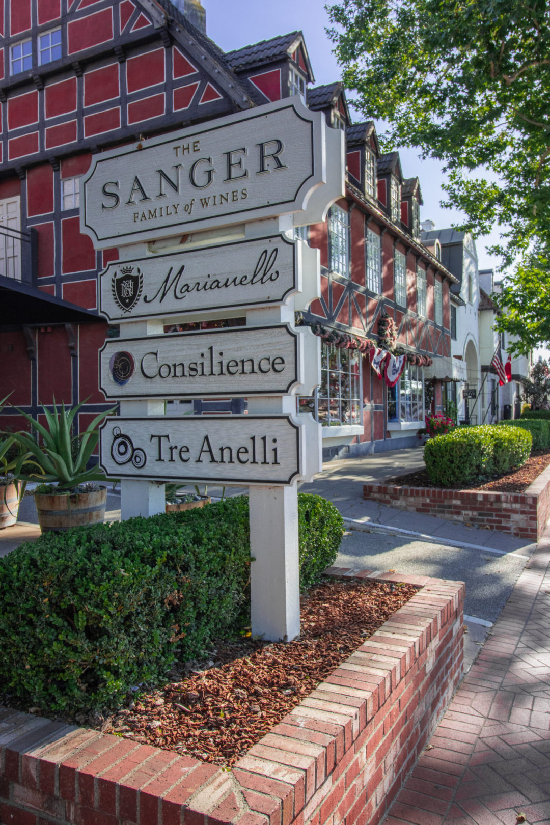A wooden sign post on Mission Drive in Solvang, California reading four local business names, one of which is "The Sanger: Family of Wines"