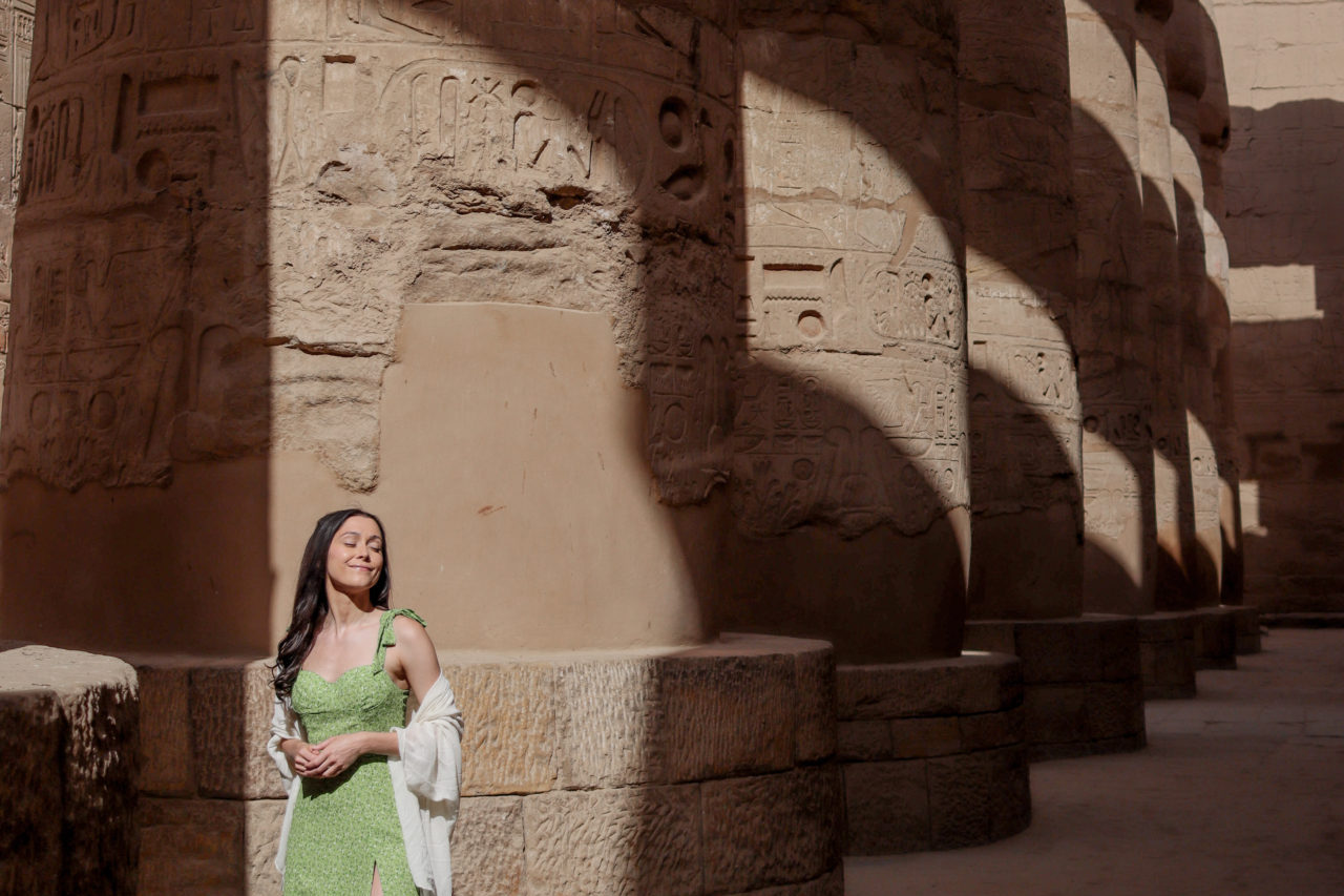 Top Ancient Sights in Egypt: Travel Blogger Jordan Gassner wearing a green dress and smiling with eyes closed while the sun peaks through the massive columns of Karnak Temple's Hypostyle Hall in Luxor, Egypt