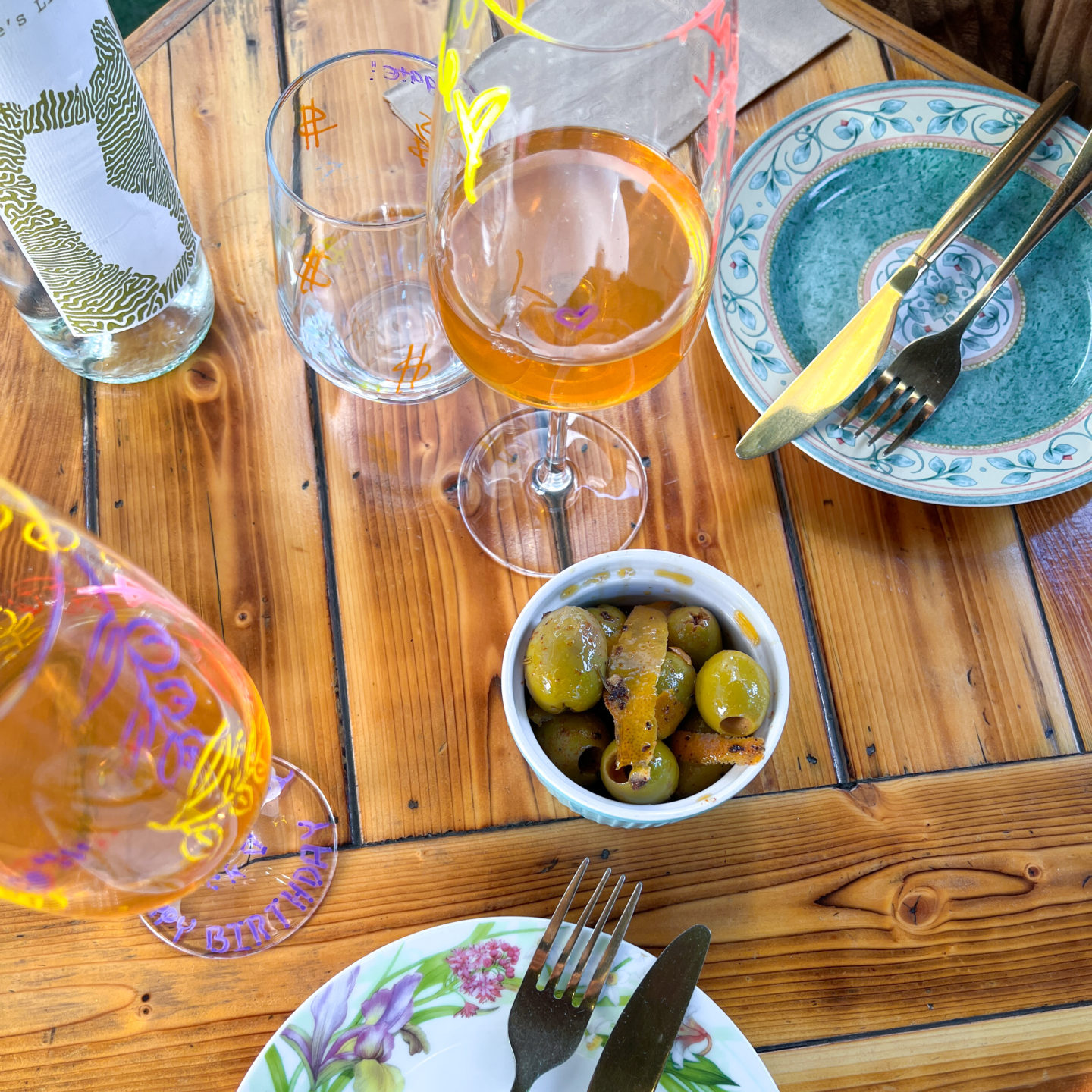 Two glasses of orange wine in funky decorated wine glasses and a plate of olive next to two floral print china plates at Good Clean Fun in Downtown LA