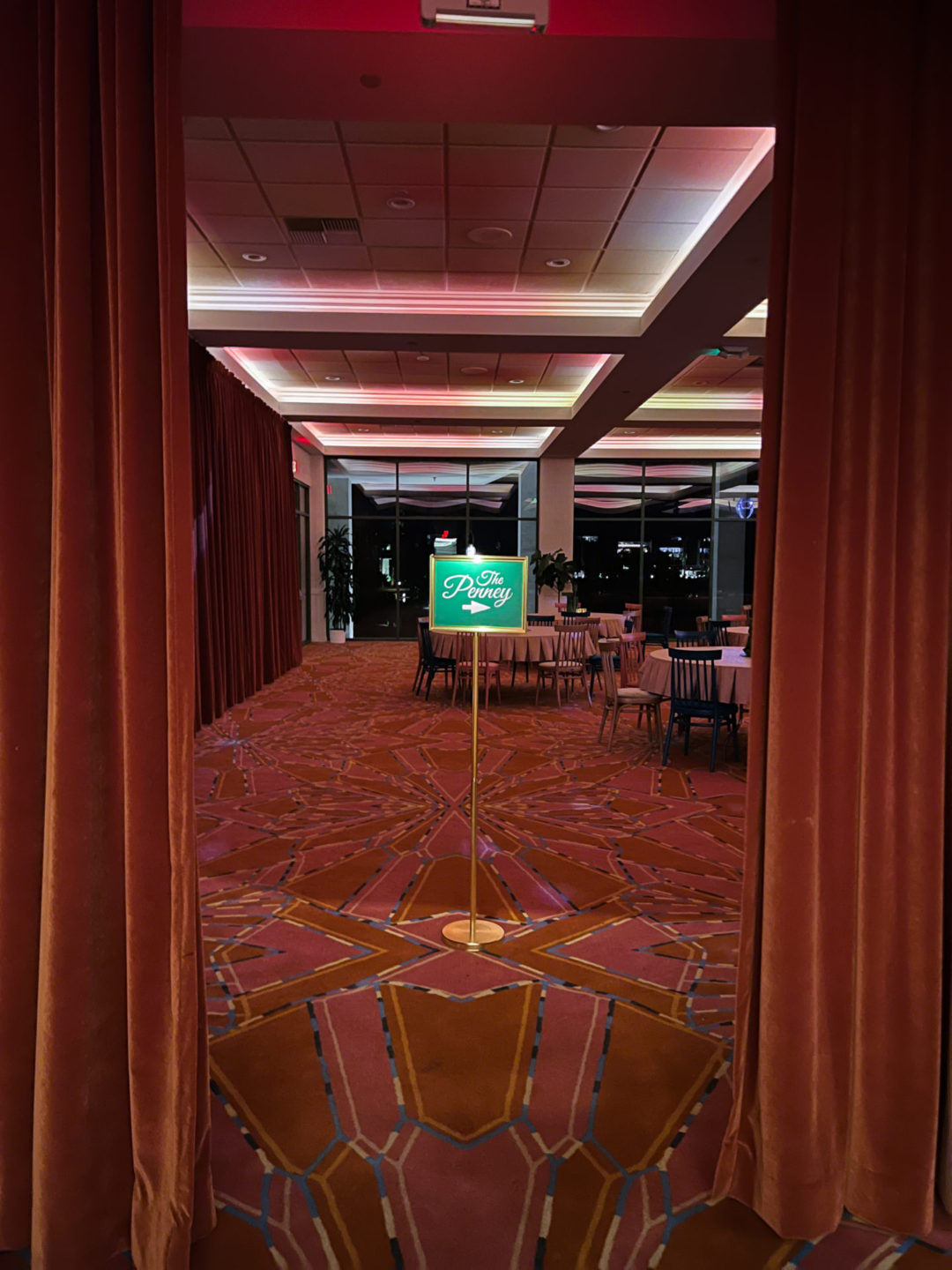 Don't Worry Darling Travel Aesthetic: A green sign with an arrow and the text "The Penny" inside a retro ballroom with red patterned floors