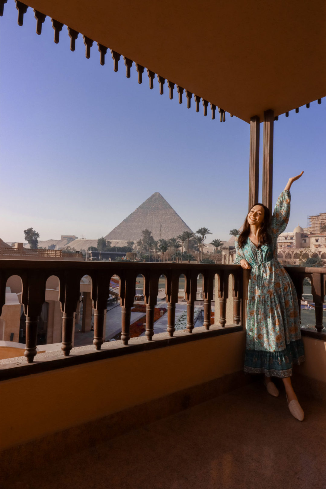 Travel Blogger Jordan Gassner leaning against a railing and smiling with her arm in the air in front of a view of the Pyramids of Giza from Marriott Mena House in Egypt
