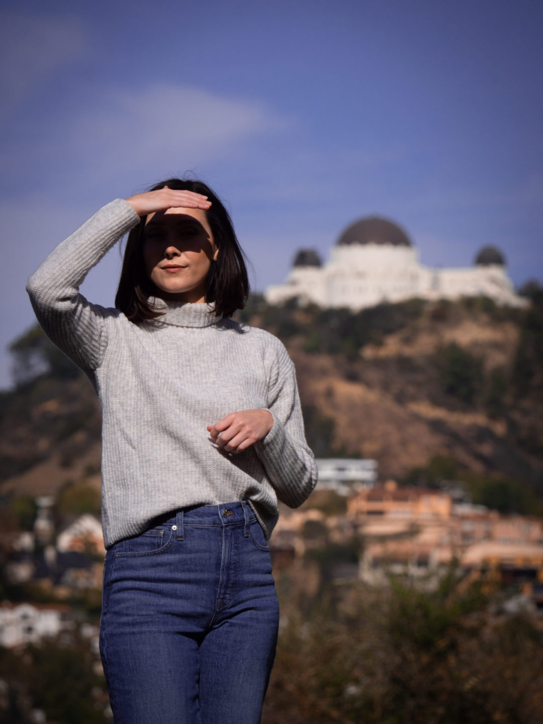 Travel Blogger Jordan Gassner shielding her eyes from the sun while standing in front of a view of LA's Griffith Observatory from Barnsdall Park