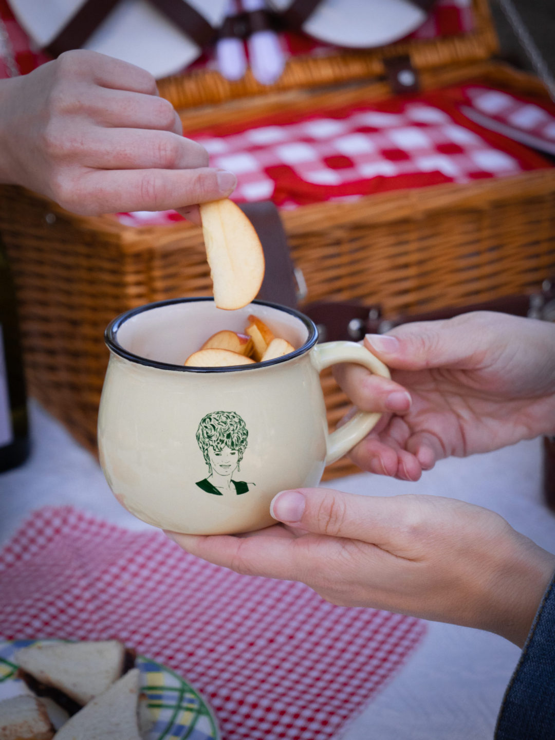 Two sets of hands enjoying sliced apples inside a Lucille's Manor branded mug during a picnic at the Noble + Proper Hotel