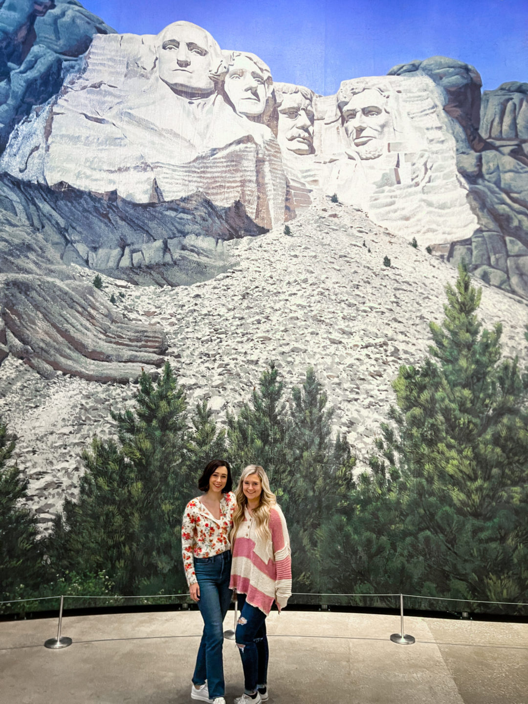Travel Blogger Jordan Gassner and her sister standing and smiling in front of the North By Northwest Mount Rushmore backdrop at the Academy Museum in Los Angeles, California