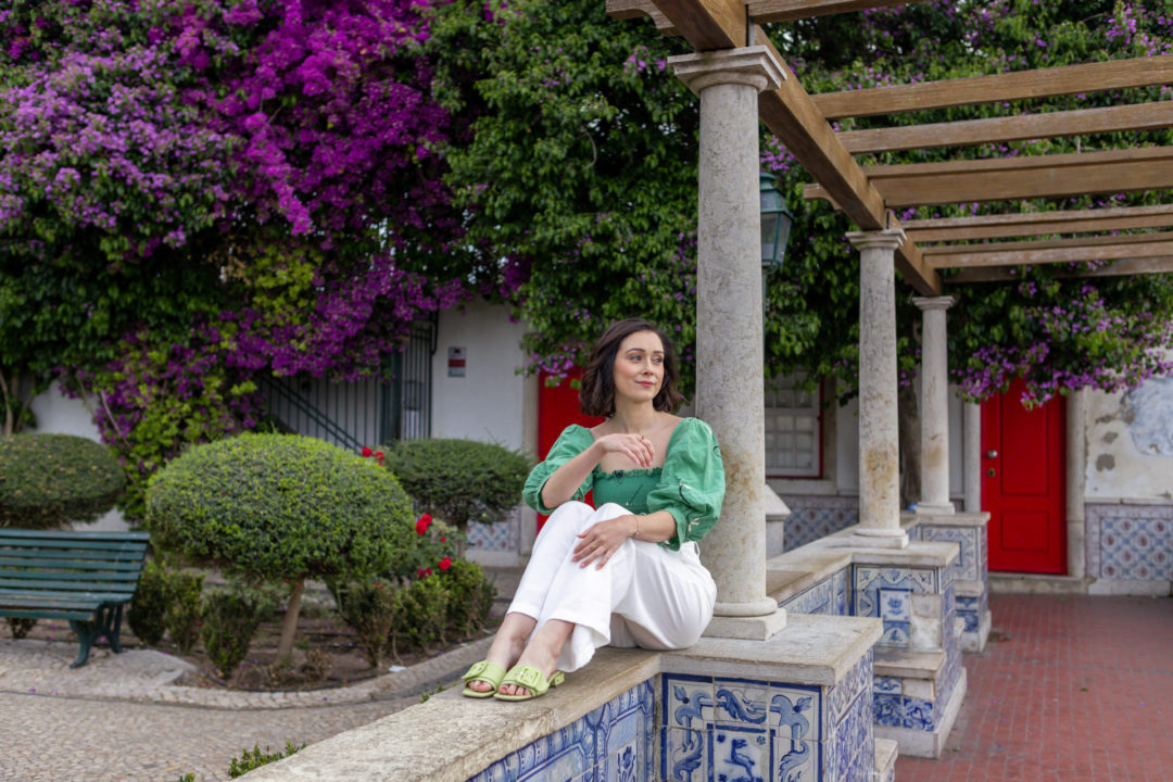 Jordan Gassner sitting on a ledge of Lisbon's Miradouro de Santa Luzia, with bright red doors, blue azulejo tiles, bougainvillea and roses in the background