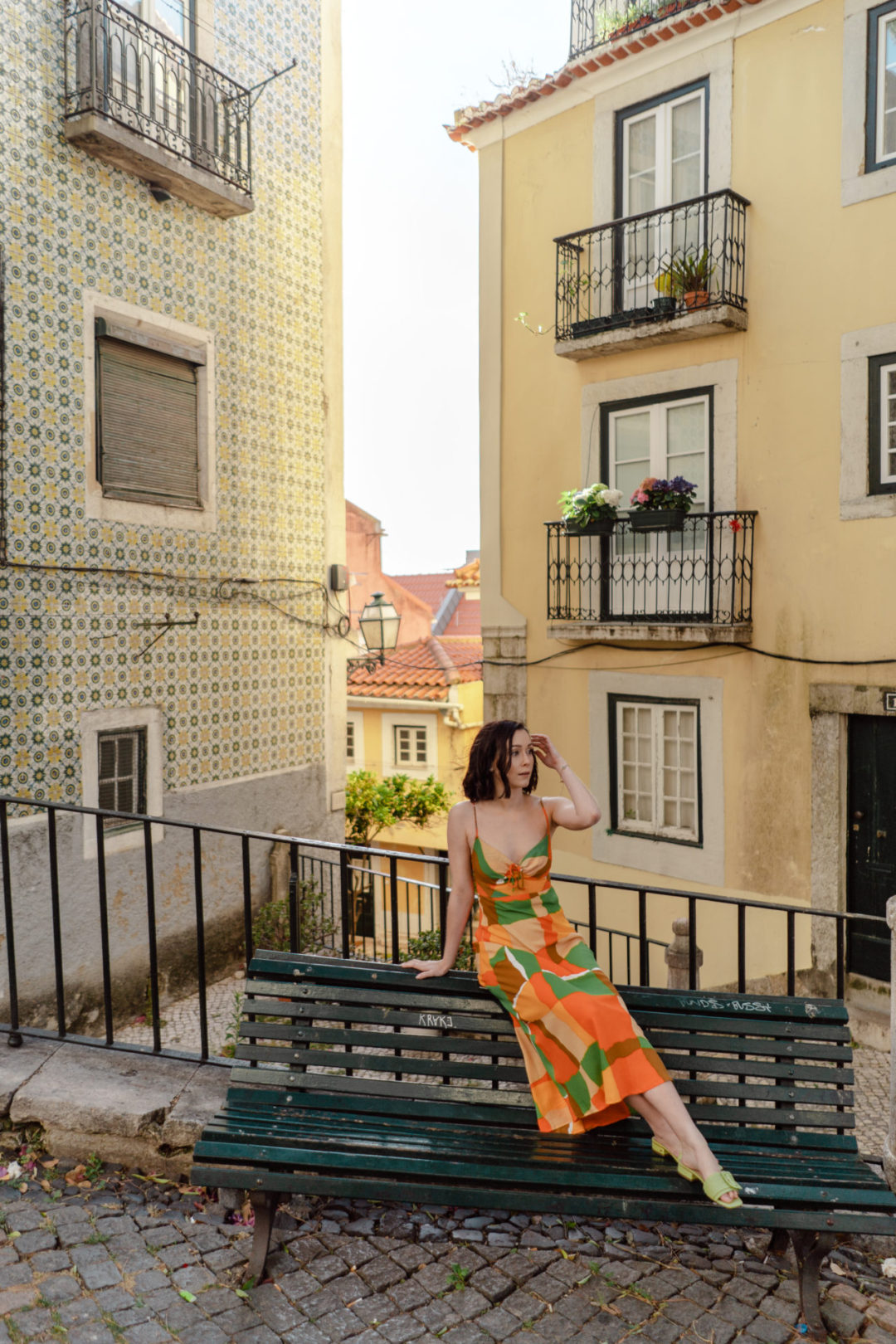 A high angle photo of Travel Blogger Jordan Gassner sitting on a green public bench in a Faithfull the Brand dress in Lisbon, Portugal