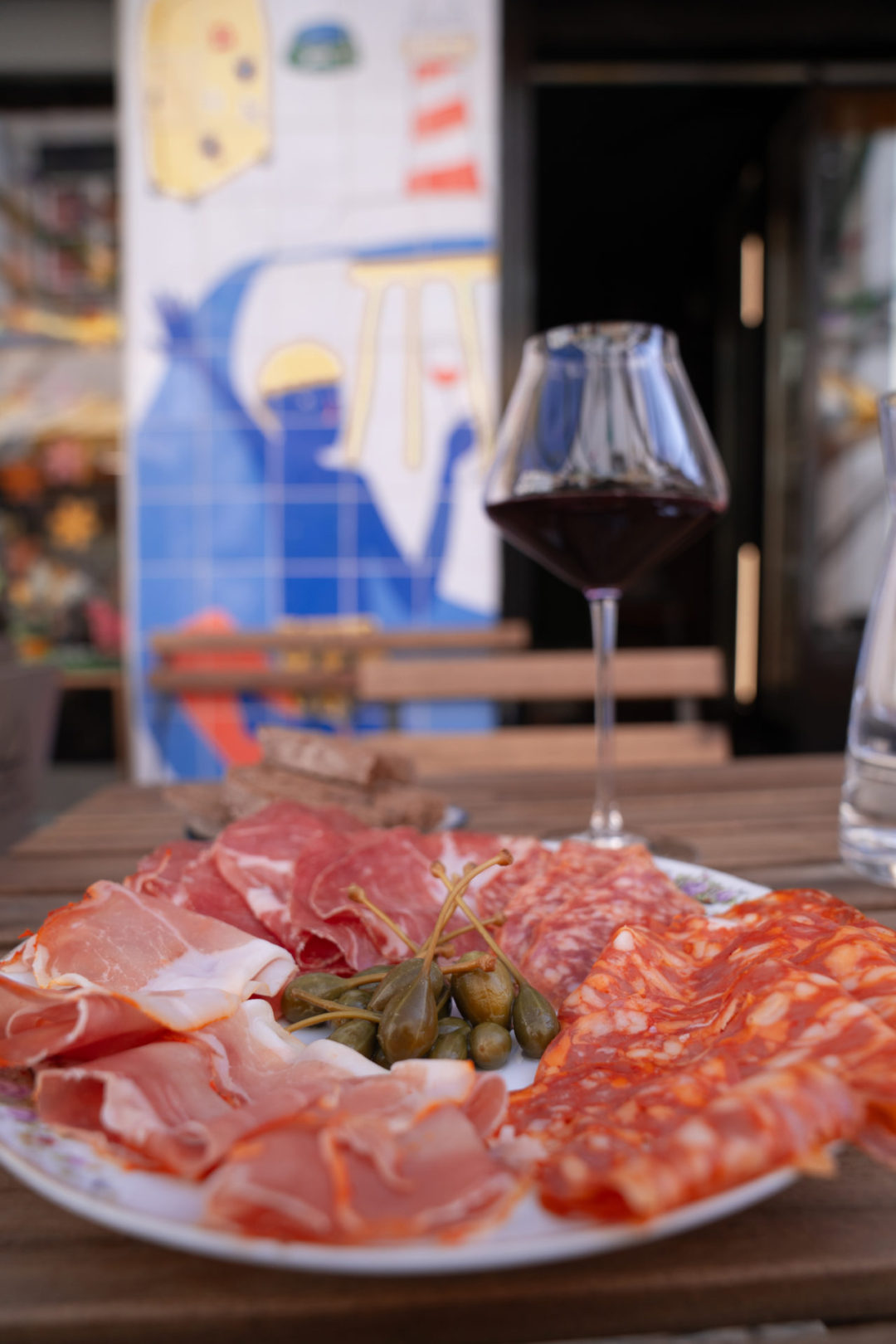 A plate of cured meats, olives and bread alongside a glass of red wine on a table outside Locals & Nomads natural wine bar in Lisbon, Portugal