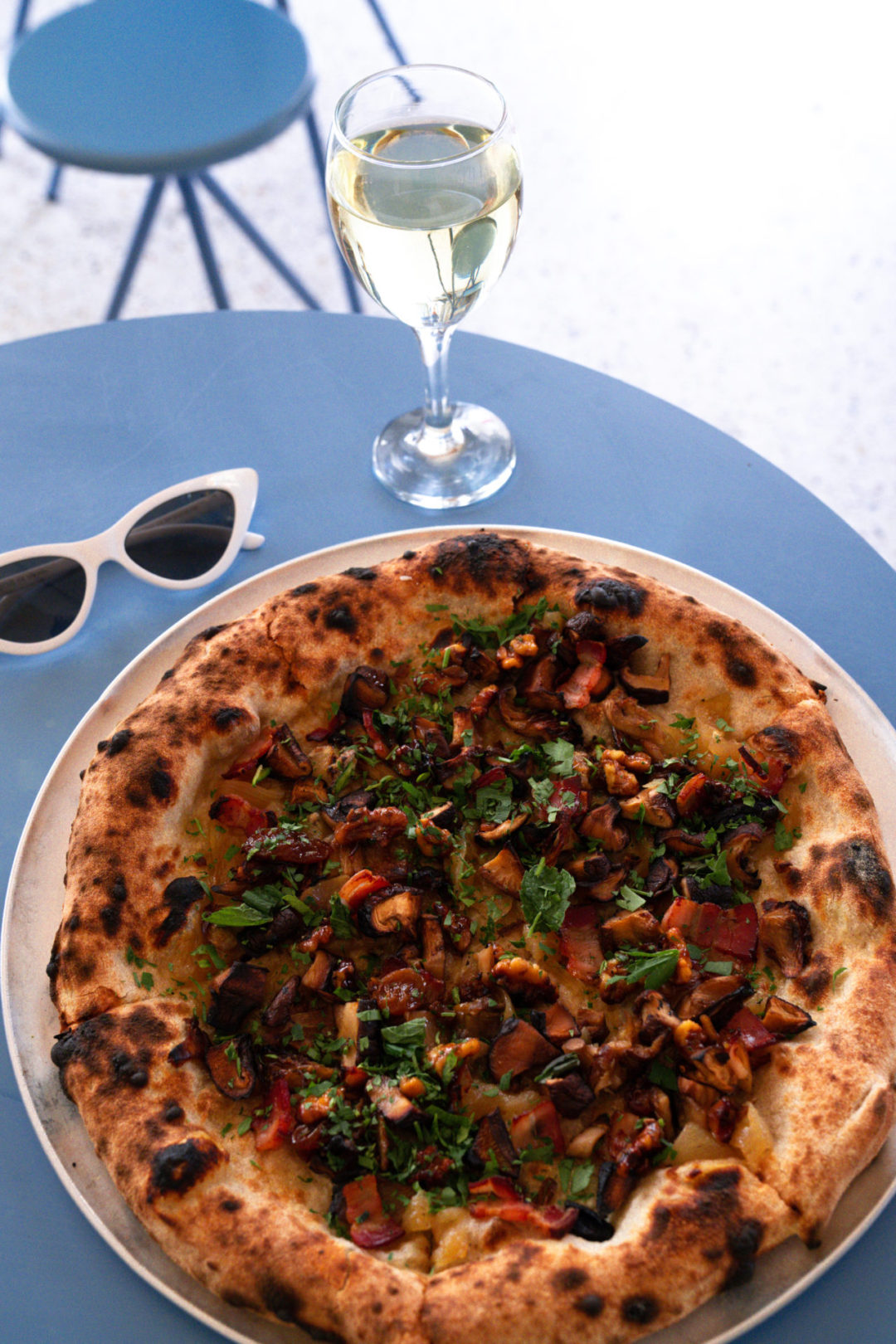 A pair of white cat eye sunglasses and a glass of white wine near a vegan mushroom pizza with mushrooms from Lupita in Lisbon