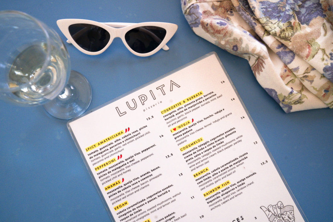 A pair of white cat eye sunglasses, a glass of white wine and Lupita's restaurant menu sitting on a blue table inside Lupita Pizzeria, one of the best Lisbon restaurants