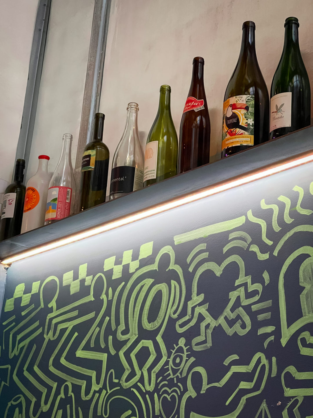 A shelf full of empty wine bottles above a wall with a green and blue mural at Tricky's in Lisbon, Portugal
