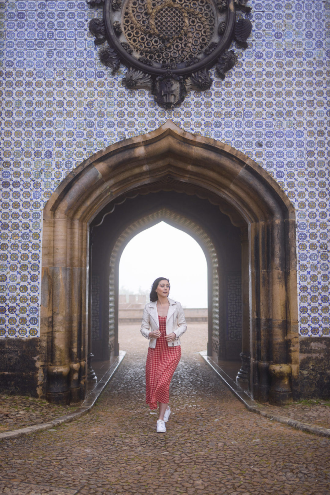 Travel Blogger Jordan Gassner walking through a misty archway while wearing a cream faux leather jacket and a MinkPink Dress at Pena Palace in Sintra, Portugal