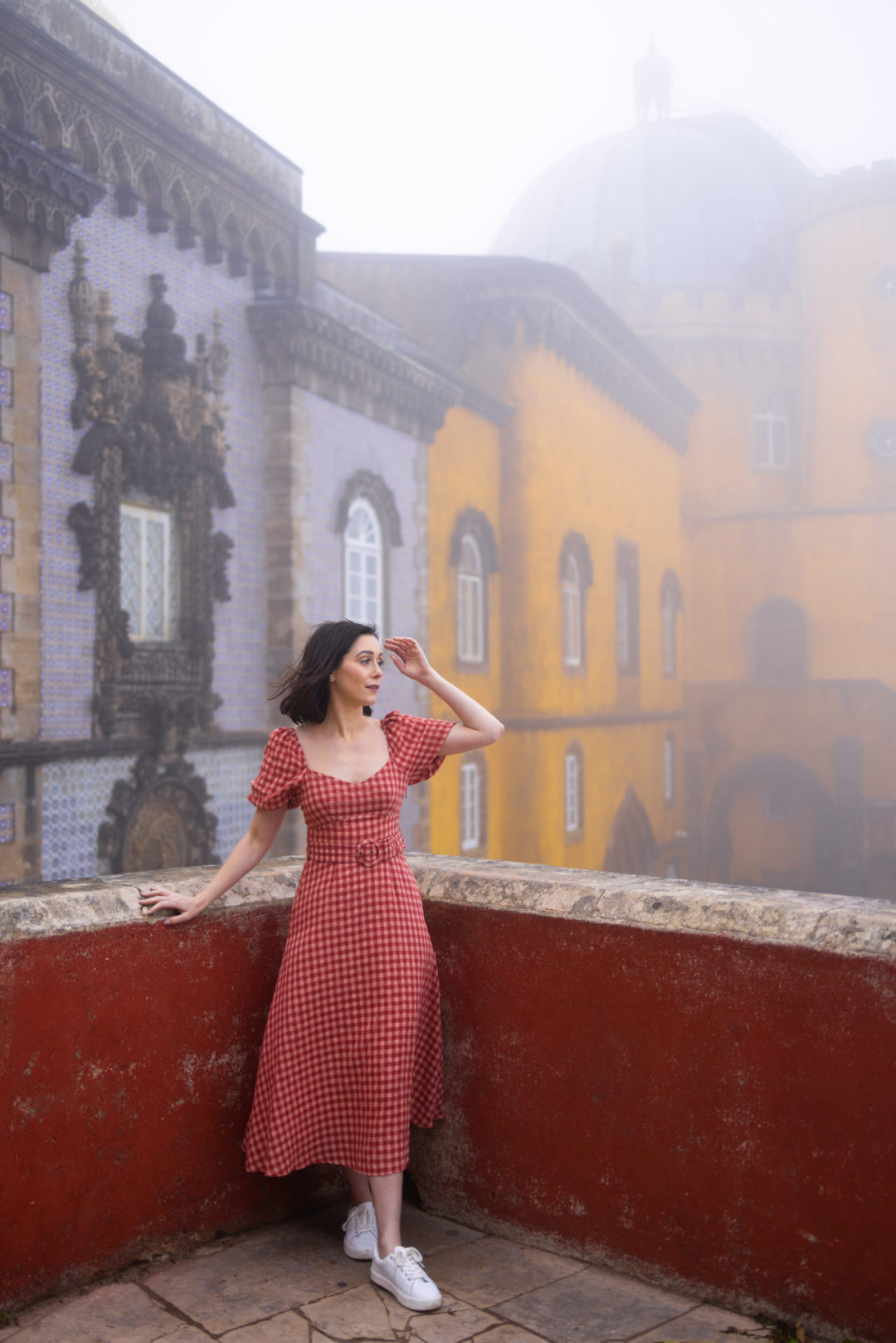 Travel Blogger Jordan Gassner fixing her hair while leaning against a red balcony in the center of the red, blue and yellow Pena Palace in Sintra, Portugal
