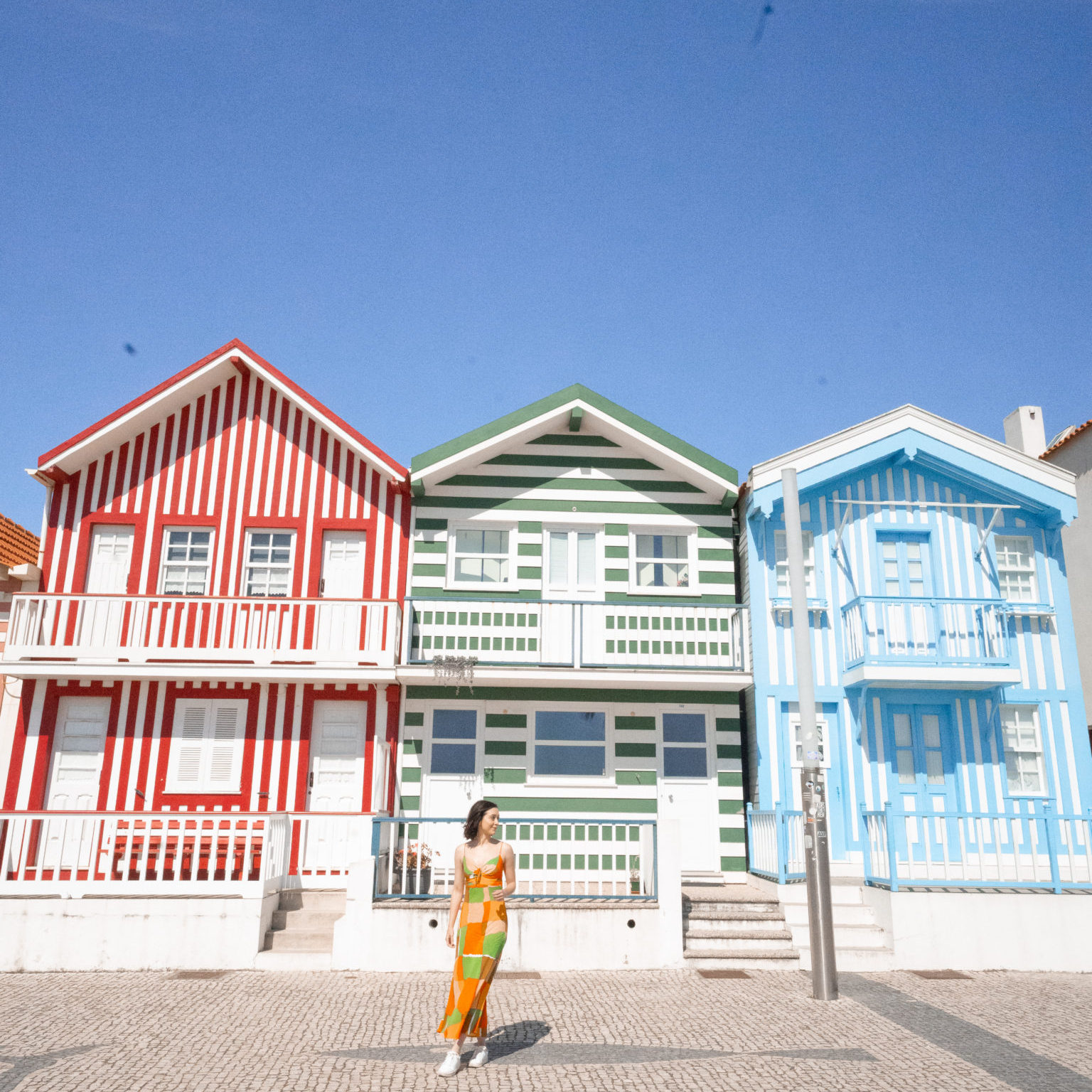 Travel Blogger Jordan Gassner wearing a bright orange and green dress in front of a row of red, green and blue striped houses in Costa Nova, Portugal